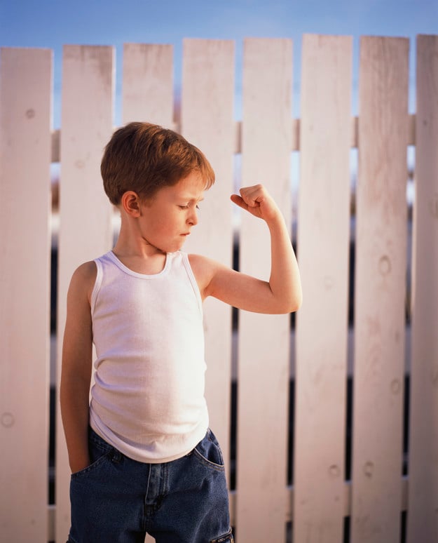 En ung gutt som flekser ene armen. Han går i mørk jeans og hvit singlet. Bak han er hvitt stakittgjerde. 