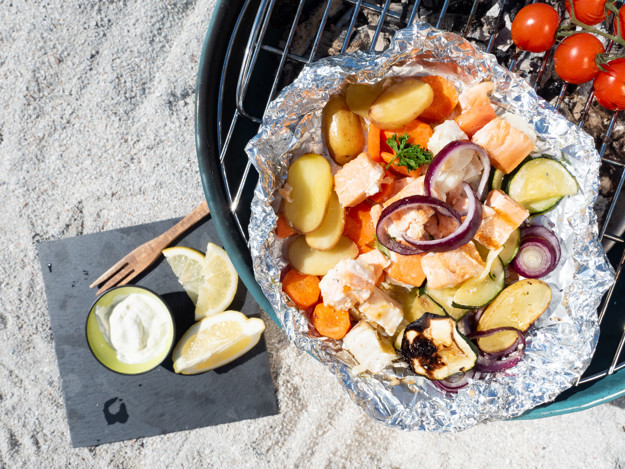 Strand med grill. Oppå grillen er aliminiumsfolie med grønnsaker og fisketerninger og tomater ved siden av.